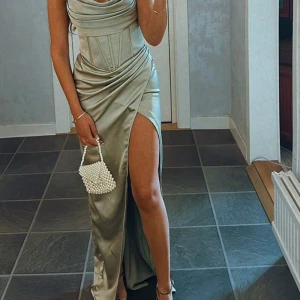 Champagnefärgad långklänning med slits - Säljer en elegant långklänning i champagnefärg med draperad överdel och markerad midja. Klänningen har en hög slits på sidan och smala axelband. Materialet är glansigt och ger en lyxig känsla, perfekt för speciella tillfällen. Snygg och trendig look!