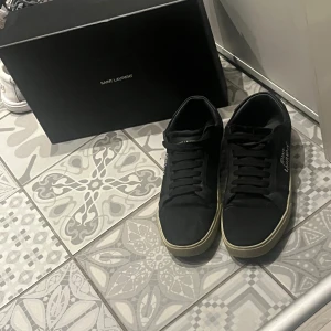 Svarta Saint Laurent sneakers - Svarta sneakers från Saint Laurent med handskriven logga på sidan och klassisk snörning. Skorna har en rund tå och platt sula i beige ton. Materialet är skinn och textil, vilket ger en lyxig och stilren vibe. Perfekt för dig som gillar designer och vill sticka ut med en clean look.