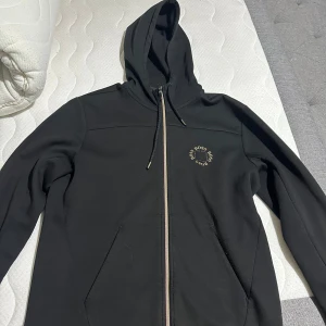 Svart hoodiejacka från Hugo Boss - Svart hoodiejacka från Hugo Boss med dragkedja framtill och gulddetaljer på bröstet. Jackan har huva med snörning och två fickor på sidorna. Perfekt för en chill och stilren look.