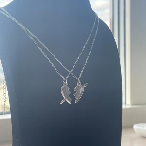 Silverhalsband med delat hjärta - Två silverfärgade halsband med matchande hängen som tillsammans bildar ett delat hjärta. Perfekt för dig och din bästa vän! Kedjorna är tunna och blanka, och hjärthängena har en cool, kantig design med små detaljer. Snyggt att bära tillsammans eller var för sig. Rostfri och kjedjan är 40cm lång