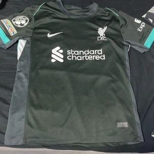 Liverpool FC svart Nike matchtröja - Liverpool FC Champions League tröja från Nike i svart med turkosa detaljer på ärmarna och sidorna. Kortärmad modell med klubbmärke och sponsortryck på bröstet, samt UEFA Foundation och Champions League-märke på ena ärmen. Tillverkad i lätt polyester för maximal komfort.