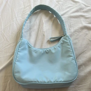 Ljusblå axelväska - Säljer en ljusblå axelväska i syntetmaterial med enkel, clean design. Väskan har en bred axelrem. Insidan är vit och har två mindre innerfack. Perfekt storlek för det viktigaste och ger en fräsch vibe till din outfit. Defekter syns på bild 3 och 4