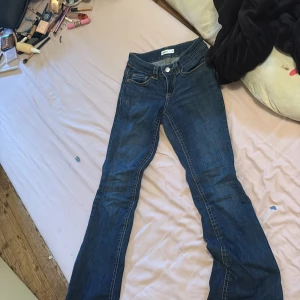 Blå bootcut jeans med fickdetaljer - Säljer ett par klassiska blå jeans med bootcut-modell och snygga fickor bak med lock och knapp. Jeansen har normal passform och är tillverkade i denim med kontrastsömmar. Perfekta för dig som gillar en tidlös look med lite utsvängda ben.