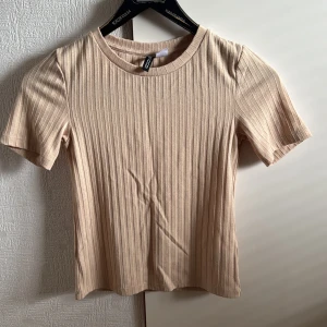 Beige ribbad topp från H&M Divided - En beige ribbad topp från H&M Divided med korta ärmar och rund halsringning. Toppen har en slim passform och är tillverkad i mjuk bomullsmix som känns skön mot huden. Perfekt att styla med jeans eller kjol för en enkel och snygg look.