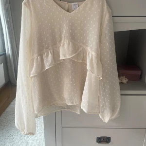 Vit prickig blus med volang från Lindex - Supersöt vit blus från Lindex med diskret prickigt mönster och volangdetaljer framtill. Blusen har långärmade, lätt puffade ärmar och är gjord i ett tunt, luftigt material som känns skönt mot huden. Perfekt för dig som gillar romantisk och feminin stil.