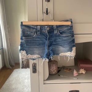 Blå jeansshorts med spets från H&M - Snygga blå jeansshorts från H&M med fransig kant och vit spetsdetalj på benen. Klassisk femficksmodell med knapp och dragkedja framtill. Perfekt för dig som gillar en chill och trendig sommarstil.