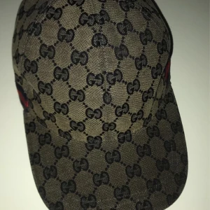 Gucci keps med GG-mönster och röd/blå detalj - Snygg keps från Gucci med klassiskt GG-monogram i svart på beige bakgrund. Kepsen har en böjd skärm och justerbar rem baktill. På sidan finns en ikonisk röd och blå rand. Tillverkad i tålig bomullscanvas, perfekt för dig som vill sticka ut.