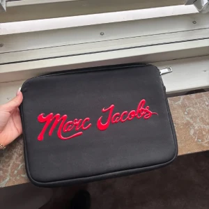 Svart datorväska Marc Jacobs - Perfekt till skolstarten! Svart datorväska från Marc Jacobs med coolt rött logotyptryck på framsidan och diskret vit text på baksidan. Väskan har en mjuk, rektangulär form och dragkedja upptill. Perfekt för att bära din laptop med stil.