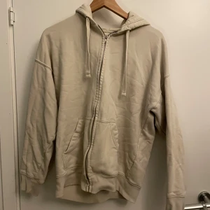 Beige hoodie med dragkedja från H&M - Säljer en beige hoodie från H&M i storlek M. Tröjan har huva med snörning, dragkedja framtill och en stor känguruficka. Materialet är mjuk bomull och passformen är loose, perfekt för chill dagar. Långärmad och enkel design som funkar till allt.