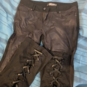 Svarta Rock Rebel byxor med snörning - Svarta byxor från Rock Rebel med glansig finish och snygga snörningsdetaljer längs benen. Byxorna har klassisk femficksdesign, dragkedja och knapp framtill. Materialet känns som en blandning av bomull och syntet, vilket ger en cool look.