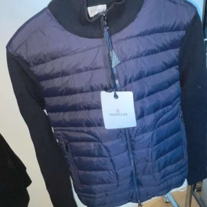 Blå Moncler Cardigan - Snygg mörkblå pufferjacka från Moncler med stickade ärmar och quiltad framsida. Jackan har dragkedja framtill, hög krage och Moncler-logga på ärmen. Perfekt för dig som vill ha en stilren och sportig look med exklusiv känsla.