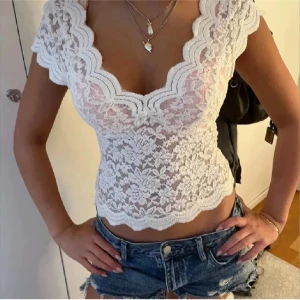 Vit spetsblus från Zara med v-ringning - Supersöt vit blus i spets från Zara med djup v-ringning och korta ärmar. Blusen har blommigt spetsmönster och vågiga kanter längs halsringningen och nederkanten. Perfekt att styla med jeans eller kjol för en trendig look.