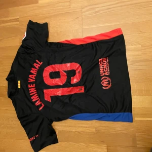 FC Barcelona Lamine Yamal tröja Nike storlek S - Snygg FC Barcelona fotbollströja med Lamine Yamal och nummer 19 på ryggen. Svart bas med röda och blå detaljer på sidorna, röd Spotify-logga på bröstet och Nike-logga. FCB-emblem i svart med blå och röd kant. Kortärmad och tillverkad i lätt polyester.