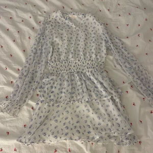 Blommig vit kortklänning Gina Tricot - Supersöt vit kortklänning från Gina Tricot med blått blommigt mönster. Klänningen har långärmade transparenta ärmar, smock i midjan och volangdetaljer på kjolen. Perfekt för dig som gillar romantisk och fräsch stil. Materialet känns lätt och luftigt.