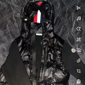 Svart pufferjacka från Moncler - Snygg svart pufferjacka från Moncler med glansig finish och stickade svarta ärmar. Jackan har huva med Monclers klassiska röd, vit och blå detalj på insidan samt dragkedja framtill. Perfekt för dig som vill ha en stilren och sportig look.