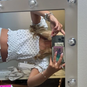 Vit prickig croppad blus  - Supersöt vit blus med svarta prickar och smockad midja. Toppen är croppad med korta puffärmar och volangdetaljer vid ärmslut och nederkant. Perfekt för dig som letar stockolmstil kläder