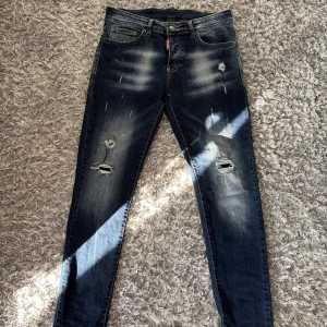 Mörkblå Dsquared2 skinny jeans - Säljer ett par mörkblå skinny jeans från Dsquared2 med slitningar och hål på benen för en riktigt edgy look. Klassisk femficksmodell med DSQ2-logga bak i midjan. Jeansen har en tight passform och är tillverkade i stretchig denim.