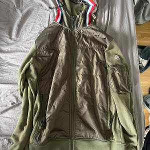 Säljer en olivgrön jacka från Moncler med dragkedja och huva. Jackan har en broderad Moncler-logga på ärmen och detaljer i rött, vitt och blått på huvan. Perfekt för kyliga dagar!