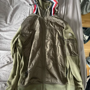 Olivgrön jacka från Moncler - Säljer en olivgrön jacka från Moncler med dragkedja och huva. Jackan har en broderad Moncler-logga på ärmen och detaljer i rött, vitt och blått på huvan. Perfekt för kyliga dagar!