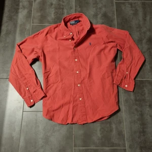 Röd Polo Ralph Lauren skjorta  - Säljer en snygg röd skjorta från Polo Ralph Lauren med klassisk krage och knappar framtill. Skjortan har en broderad logotyp i blått på bröstet och långa ärmar med knappar vid ärmsluten. Perfekt för en stilren look!
