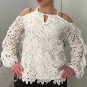 Vit spetsblus  - Säljer en elegant vit blus med vackra spetsdetaljer och offshoulder-design. Blusen har långa ärmar med volanger och ett blommigt mönster. Perfekt för en stilfull look.
