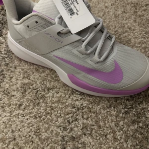 Nike Vapor grå och lila sneakers - Snygga Nike Vapor sneakers i grått med lila detaljer. Skorna har en modern design med snörning och en bekväm sula. Perfekta för både träning och vardag. Köpta för ca 1700 kr ett par månader sedan helt oanvända med box osv. Riktigt sköna skor i storlek 44 passar även 43 sitter riktigt bra 