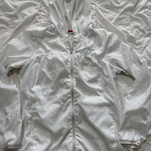 Vit vindjacka med huva - Säljer en stilren vit vindjacka med huva och dragkedja framtill. Jackan har långa ärmar och en liten logga i turkos på bröstet. Perfekt för blåsiga dagar!