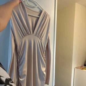 Champagnefärgad långärmad klänning - Elegant champagnefärgad klänning med långa ärmar och v-ringad design. Tillverkad i ett glansigt material som ger en lyxig känsla. Perfekt för speciella tillfällen.