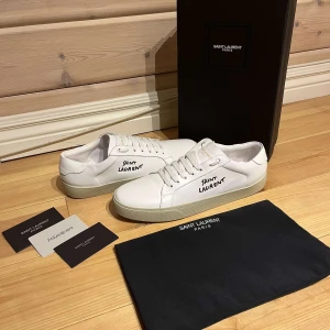 Saint Laurent sneakers vita - Snygga vita sneakers från Saint Laurent med svart logotyp på sidan. Skorna har en klassisk design med snörning och en bekväm gummisula. Perfekta för en stilren look.