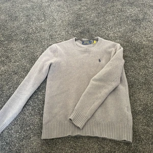 Grå tröja från Polo Ralph Lauren - Säljer en stilren grå tröja från Polo Ralph Lauren. Tröjan är tillverkad i ett mjukt material och har långa ärmar samt ribbade muddar vid ärmslut och nederkant. Perfekt för en avslappnad och elegant look.
