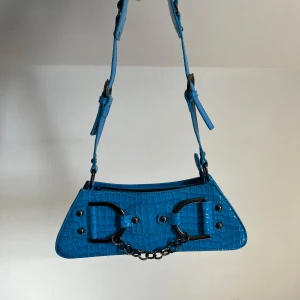 Blå axelväska från NA-KD - Snygg blå axelväska med krokodilpräglat mönster och en cool kedjedetalj framtill. Väskan har justerbar axelrem och stängs med dragkedja. Perfekt för att ge din outfit en färgklick!