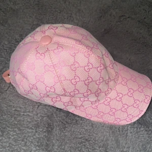 Rosa keps från Gucci - Snygg rosa keps från Gucci med deras ikoniska mönster. Kepsen har en justerbar rem baktill och är perfekt för att ge din outfit en lyxig touch. Den är tillverkad i ett slitstarkt material och har en klassisk design.