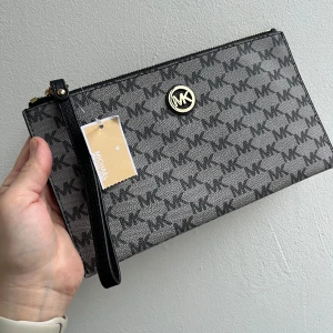 Svart och grå kuvertväska från Michael Kors - Snygg kuvertväska från Michael Kors i svart och grått med det ikoniska MK-mönstret. Väskan har en dragkedja upptill och en praktisk handledsrem. Perfekt för att hålla dina nödvändigheter säkra och stiliga. Köpt i Usa