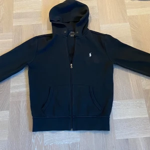 Svart huvtröja från Polo Ralph Lauren - Säljer en stilren svart huvtröja från Polo Ralph Lauren med dragkedja och broderad logga på bröstet. Tröjan har långa ärmar och en praktisk huva. Perfekt för en avslappnad look. Storlek S och passar dig mellan 170-180! Tröjan är använd lite grann men fortfarande ett fint skick!💯Nypris 1700, mitt pris 549 kr👍 Vid frågor eller om man vill ha fler bilder eller liknande så är det bara att höra av sig!! //Leon😊