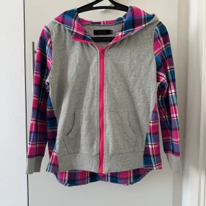 Grå och rutig hoodie med dragkedja - Säljer en cool hoodie med grå framsida och färgglatt rutigt mönster i rosa, blått och lila på ärmar och baksida. Har storlek S, men passa som Xs