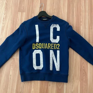 Blå sweatshirt från Dsquared2 - Säljer en snygg blå sweatshirt från Dsquared2 med stor text 'ICON' i vitt och 'DSQUARED2' i gult på framsidan. Tröjan har en klassisk rund hals och långa ärmar. Perfekt för en avslappnad stil. Storlek 12y motsvarar XS