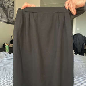 Svart Kjol - Yves Saint Laurent - Elegant svart kjol från Yves Saint Laurent i 100% ull. Kjolen har en klassisk design med en rak passform och diskreta veck framtill. Perfekt för en stilren look.