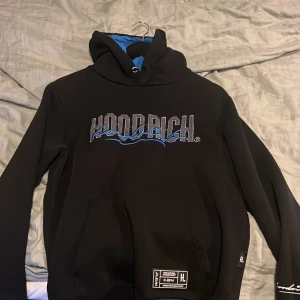 Svart hoodie från Hoodrich - Säljer en svart hoodie från Hoodrich med blå och grå detaljer. Den har en stor logga på framsidan och en känguruficka. Perfekt för en avslappnad stil. 