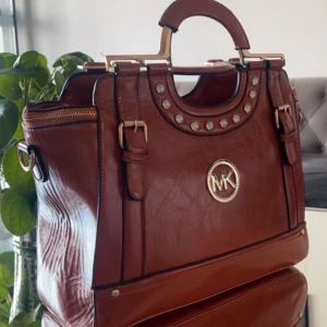 Brun handväska från Michael Kors - Snygg brun handväska från Michael Kors med metallogga framtill. Väskan har dekorativa nitar och ett praktiskt handtag. Perfekt för att bära dina dagliga nödvändigheter med stil.