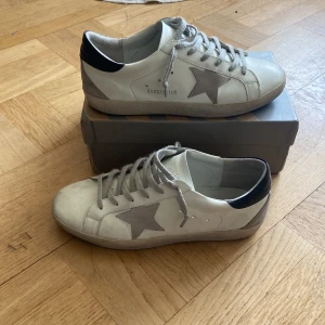 Golden goose skor - Snygga vita sneakers från Golden Goose med en grå stjärna på sidan och svarta detaljer vid hälen. Skorna har snörning och en rund tå, vilket ger en klassisk look. Perfekta för en stilren vardagsstil.
