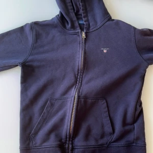 Mörkblå hoodie från Gant - Säljer en snygg mörkblå hoodie från Gant med dragkedja och huva. Tröjan har en liten broderad logga på bröstet och praktiska fickor framtill. Perfekt för en avslappnad stil.