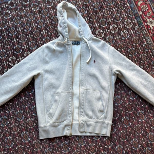 Grå hoodie från Ralph Lauren - Säljer en stilren grå hoodie från Ralph Lauren med dragkedja och huva. Tröjan har långa ärmar och en liten broderad logga på bröstet. Perfekt för en avslappnad look. Var uppmärksam om att tröjan har en liten fläck vid dragkedjan
