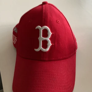 Röd keps med broderad logga - Snygg röd keps från New Era med en stor vit broderad bokstav på framsidan. Sidan har broderade detaljer med texten 'Boston' och '1901' samt en logga med strumpor. Justerbar rem baktill för perfekt passform.