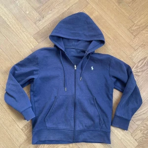 Mörkblå hoodie från Polo Ralph Lauren - Säljer en snygg mörkblå hoodie från Polo Ralph Lauren med dragkedja och klassisk logga på bröstet. Perfekt för en avslappnad stil. Den har en justerbar huva och praktiska fickor framtill.
