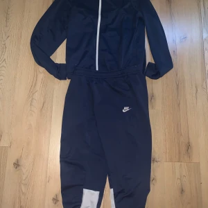 Blå träningsset från Nike - Säljer ett stilrent blå träningsset från Nike med långärmad överdel och matchande byxor. Setet har en dragkedja framtill och Nike-logga på byxan. Perfekt för träning eller en avslappnad dag.