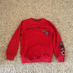 Röd tröja från Supply & Demand - Säljer en cool röd tröja från Supply & Demand med tryck och patches på bröstet och ärmen. Tröjan har långa ärmar och ribbade muddar. Perfekt för en avslappnad stil.