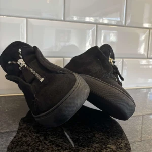 Svarta sneakers från Axel Arigato - Snygga svarta sneakers från Axel Arigato i mocka med dragkedjedetaljer. Skorna har en rund tå och en stilren design som passar perfekt för en modern look. Box plus dustbag ingår. Kom priv för minsta lilla. 