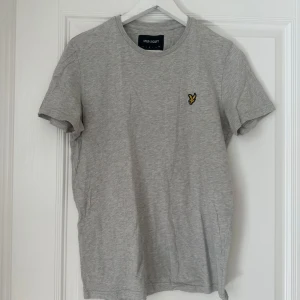 Grå t-shirt från Lyle & Scott - Säljer en stilren grå t-shirt från Lyle & Scott med ett broderat gult emblem på bröstet. T-shirten är kortärmad och tillverkad i mjuk bomull. Perfekt för en avslappnad look.
