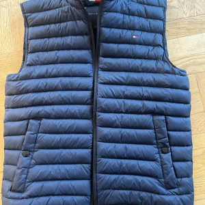 Blå quiltad väst från Tommy Hilfiger - Säljer en snygg blå quiltad väst från Tommy Hilfiger. Västen har en dragkedja framtill och praktiska fickor med knappar.  Använd ca 1-2 ggr, som ny. 