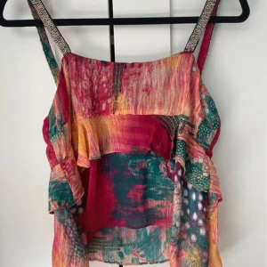 Färgglad volangtop med mönster - Säljer en färgglad linnetopp med volangdetaljer och breda axelband. Toppen har ett mixat mönster i rött, grönt, gult och inslag av vitt. Perfekt för varma dagar och ger en bohemisk känsla. skriv privat för fler frågor eller bilder, kan även gå ner i pris💞
